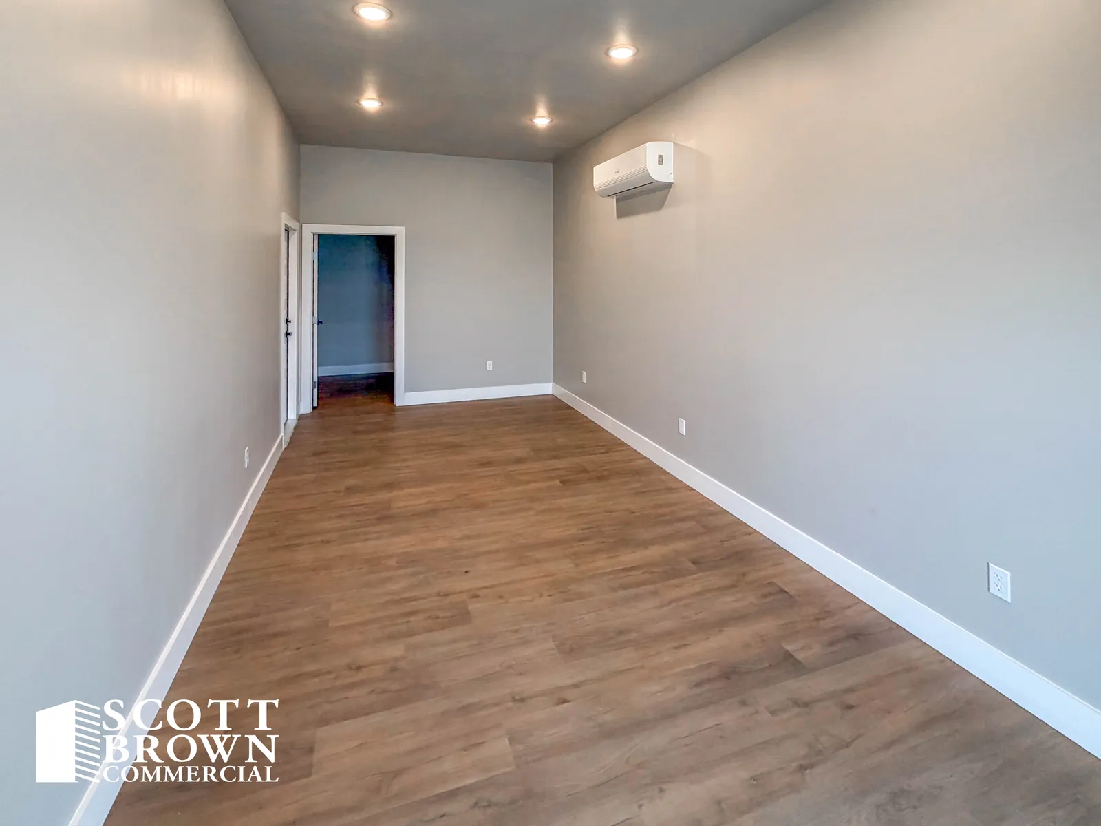 A finished office inside a Fusion Business Park suite: grey painted walls, wood-effect vinyl plank flooring, recessed downlights, a wall-mounted air conditioning head, and an open doorway through to the next room.