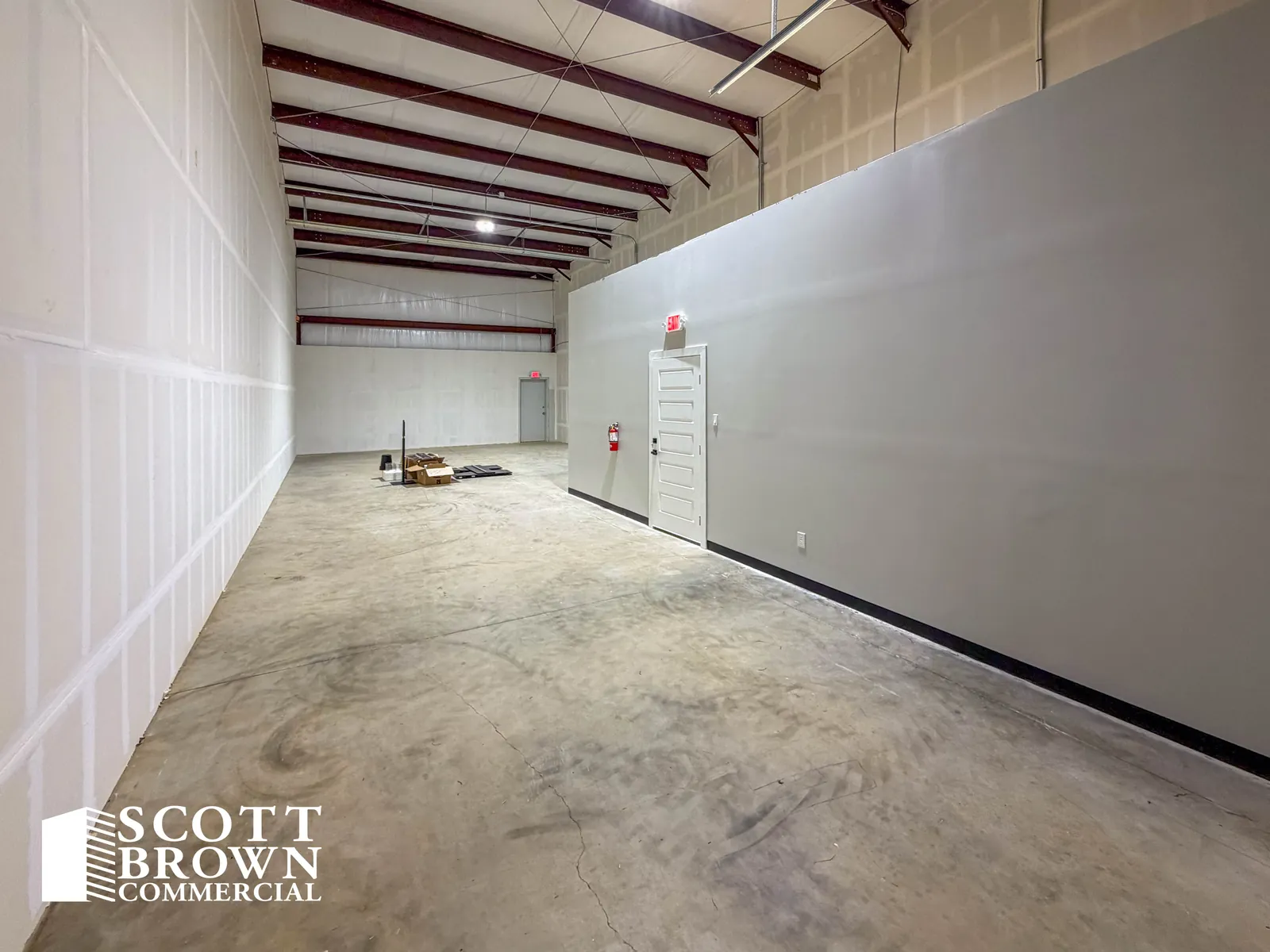 A warehouse bay at Fusion Business Park looking along the grey office partition, with red steel portal frames, white insulated metal walls, a sealed concrete floor, and a door through to the office.