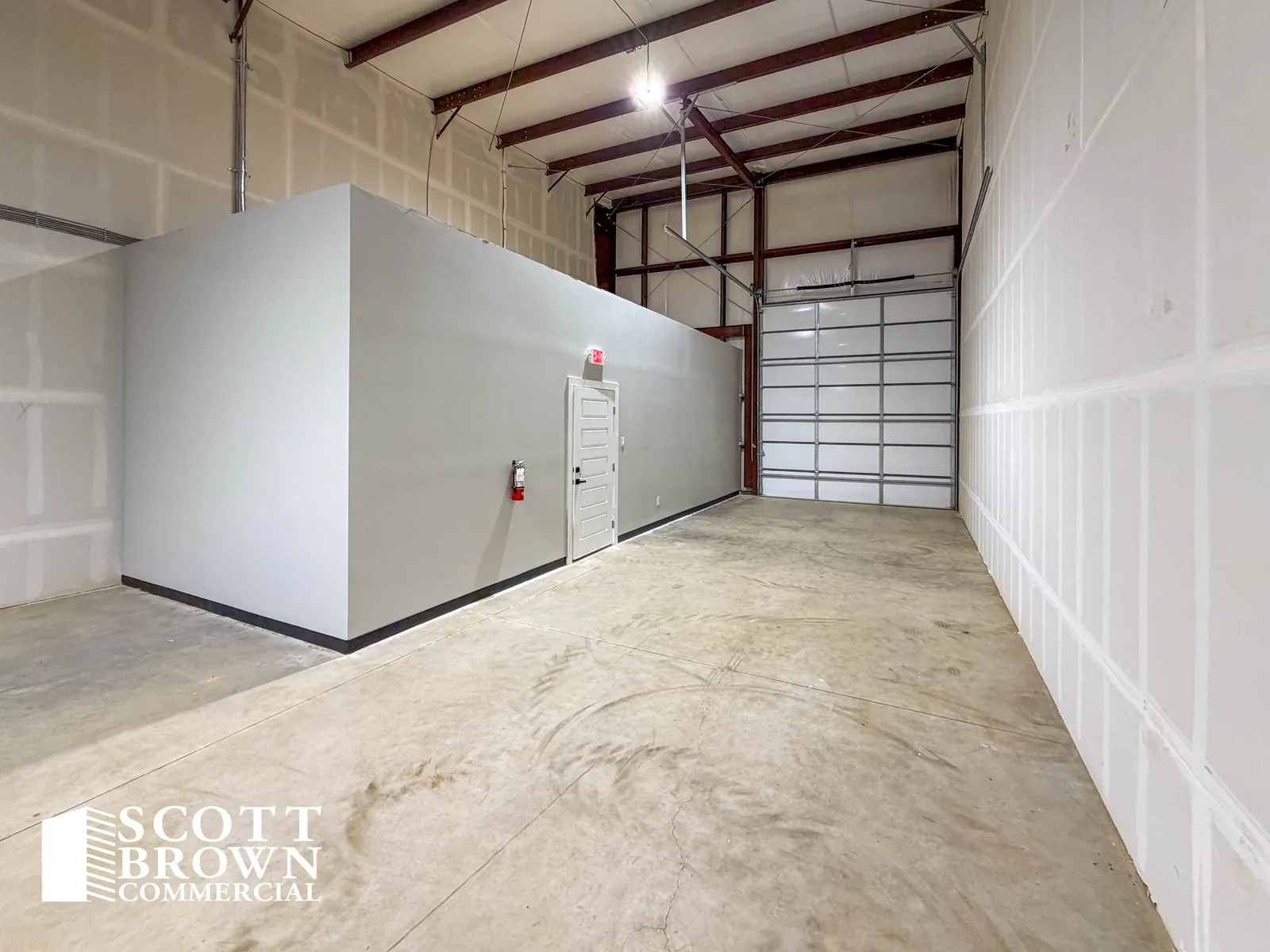 Inside a warehouse bay at Fusion Business Park: the boxed-out office block finished in grey, red steel portal frames overhead, white insulated wall lining, a sealed concrete floor, and a closed grade-level overhead door at the end of the bay.